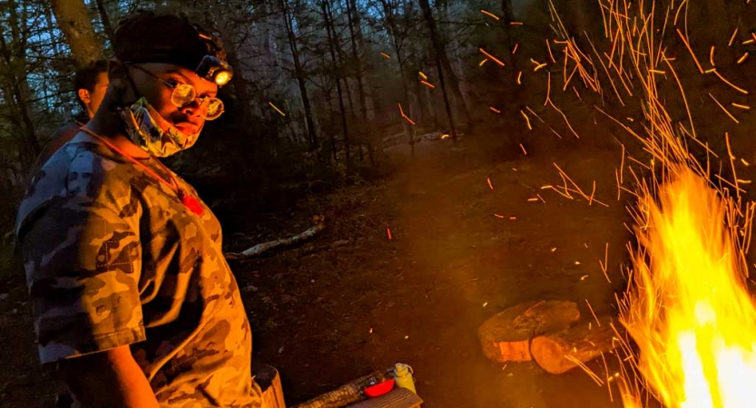 a student stands beside a campfire on a backpacking and rock climbing trip with outward bound